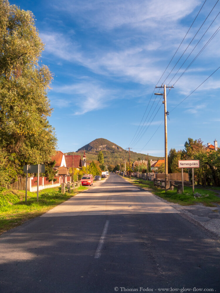 Festői falusi út Nemesgulácson, házakkal és fákkal szegélyezve. A háttérben egy domb magasodik a tiszta kék ég alatt. Villamos vezetékek futnak az út mellett.